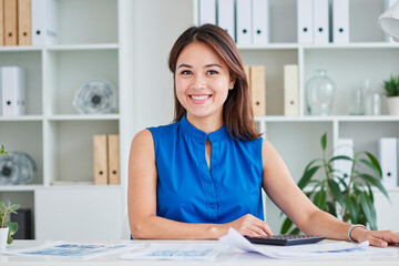 Wall Mural - Portrait of young adult Asian woman smiling while sitting at desk, working with documents and calculator, office shelves with folders and plants in background, looking into camera