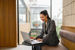 © amnaj - Smiling businesswoman working on laptop, enjoying comfortable workspace in modern office lobby with natural light