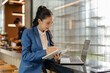 © amnaj - Asian businesswoman writing notes on her agenda using a laptop in a modern office with glass bricks wall and a bar in the background