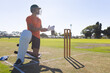 © Wavebreak Media - Cricketer in green helmet kneeling grass pitch adjusting glove strap by wooden stumps