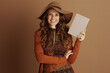 © Alliance - A relaxed woman in autumn fashion, featuring a wide-brimmed hat and orange cardigan, smiles directly at the camera while holding a pink book against a soft brown background.