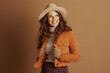 © Alliance - A confident woman with curly brown hair, wearing a beige fedora, turtleneck, and an orange suede jacket, poses against a plain background. Her stylish autumn ensemble is captured in a medium shot.