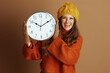© Alliance - A smiling young woman in a yellow beanie and orange turtleneck sweater holds a large wall clock against a light brown background. The scene conveys a cheerful, cozy, and seasonal mood.