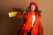 © Alliance - A cheerful adult woman in red beret, orange scarf, and orange trench coat smiles broadly at camera. She holds two patterned shopping bags against light brown backdrop, reflecting joy and autumn style