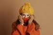 © Alliance - Woman in yellow beanie and orange ribbed sweater playfully pulls collar over lower face, revealing only eyes. Her warm gaze against plain brown background captures cozy, cheerful cold-weather mood.