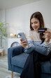 © PHOTO4U - A young woman sitting on a sofa at home smiling while holding a smartphone in one hand and a credit card in the other, representing online payment, digital finance, and e-commerce lifestyle.