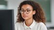 © 69 - Focused young woman with curly hair wearing glasses, working on a computer in a bright office environment, concentrated on task at hand, professional setting