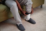 Black man sitting on sofa adjusting electronic ankle monitor on leg wearing slippers and sweatpants, concept of house arrest or home confinement with monitoring device visible