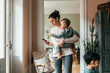 © Maskot - Woman wearing wireless in-ear headphones standing with baby boy while using smart phone at home