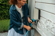 © Maskot - Smiling woman plugging electric plug in outlet on outside wall in back yard