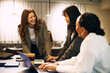 © Maskot - Happy female business professional talking with coworkers while leaning on desk in office