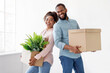 © Prostock-studio - Smiling young african american husband and wife holding cardboard boxes with plant in pot, on white wall background. Sweet home, moving to new apartment, married couple is relocation in own flat