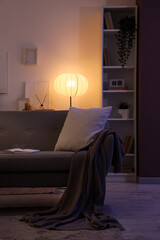 Poster - interior of dark living room with glowing lamp, sofa and shelf unit in evening