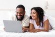 © Prostock-studio - Glad smiling loving millennial black male and female lie on bed and watch video on laptop in bedroom interior, enjoy spare time and chatting. Social networks, online call, advertising and good offer