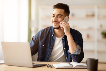 Middle-eastern bearded businessman working online from home while coronavirus pandemic, using laptop and having conversation on mobile phone, sitting at workdesk in living room, copy space