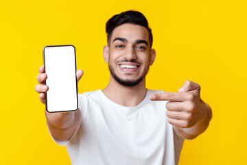 Happy handsome middle-eastern young man showing modern smartphone with empty screen, recommending nice mobile application or showing amazing offer over yellow studio background, mockup
