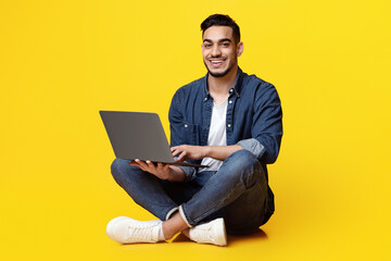 Happy arab guy in casual freelancer using modern laptop, working online, yellow studio background. Cheerful middle eastern young man independent contractor sitting on floor with notebook, copy space