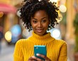 © Dadak - A young woman with curly black hair, wearing a vibrant yellow sweater, attentively looks at her teal colored cell phone outdoors.