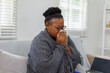 © David - African American woman wrapped on a couch,in a blanket, with flu virus blowing her nose at home.She appears to be sick or to have allergies.
