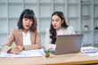 © amnaj - Two young businesswomen working together, analyzing financial charts and data on a laptop, discussing strategies and collaborating on a project in a modern office environment