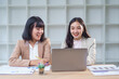 © amnaj - Two asian businesswomen discussing, brainstorming and working together using laptop in office meeting analyzing financial chart documents