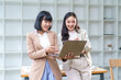 © amnaj - Two asian businesswomen discussing over laptop in modern office, smiling and collaborating on a project