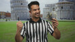 Young hispanic man in referee uniform holding us dollars with pisa tower in the background, smiling joyfully in a city street setting.