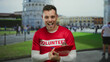 Young man in red volunteer shirt holds clipboard with pisa tower in background demonstrating cheerful urban community involvement.