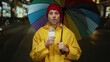 © Krakenimages.com - Young man reporter holding microphone under colorful umbrella at night street wearing yellow raincoat and red beanie.