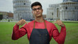 Young man wearing glasses and apron gestures in front of iconic leaning tower in lively outdoor pisa setting.
