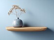 © Arwin - A light blue wall background showcases a simple wooden shelf holding a decorative, speckled gray vase with dried flowers.