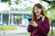 © maeching - Asian female student in warm clothes using phone to chat with friends and ready for video call while in university.