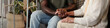© pressmaster - Young Black man holding hands with senior Caucasian woman sitting together indoors, showing support and compassion, partial view with walking aids and plant in background