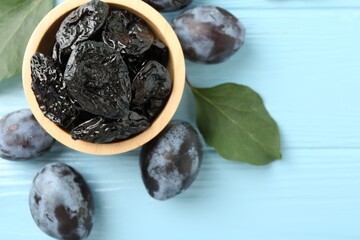 Wall Mural - Dried prunes in bowl, leaves and fresh plums on light blue wooden table, flat lay. Space for text