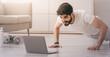 © Prostock-studio - New reality during covid-19 pandemic, home gym and online workout. Handsome arabian bearded young man doing push-ups in living room interior, looking at laptop with dumbbells, bottle of water on floor
