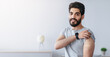 © Prostock-studio - Recommendations for vaccination and immunization of population, health care concept. Cheerful arabian bearded young male shows hand with band aid after injection in living room interior, empty space
