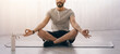 © Prostock-studio - Meditation and yoga at home, self-isolation. Cheerful arabian bearded millennial man sits on mat in lotus position, rest on floor with bottle of water in living room interior on window background