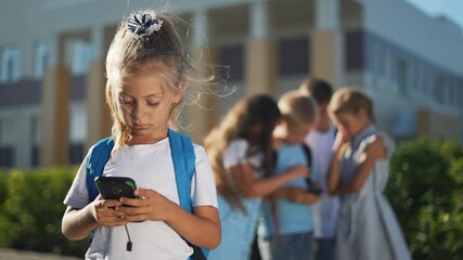 Girl using smartphone while peer group gathers near school playground showing child looking at screen and whispering suggesting bullying or exclusion with visible backpack and focused face and serious