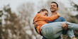 © qunica.com - A cheerful young couple enjoys a playful moment together in nature. The man lifts the smiling woman in his arms, both displaying happiness and a sense of togetherness on a serene outdoor day.