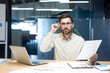 © Tetiana - Portrait of a young man working in an office with documents, sitting at a table, supporting his glasses with his hand, looking at the camera