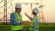© Victoriia - Two energy engineers work with computer tablet outdoors. Engineers in helmet, on field with electric towers, teamwork. Business people, power construction. Electrical with high voltage pylon, sunset.