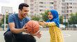 © Mono - Happy father and daughter playing basketball on an outdoor court, with the girl wearing a blue hijab and yellow dress.