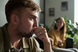 © pressmaster - Caucasian young adult man sitting indoors holding hand to mouth appearing thoughtful, while blurred Caucasian young adult woman sitting gesturing with hands during couple psychotherapy session