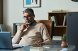 © pressmaster - Middle aged South Asian man sitting at desk working on laptop, wearing eyeglasses and wireless earphones, resting chin on hand, appearing focused in modern office setting