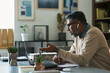 © pressmaster - Middle aged Indian man with gray beard sitting at desk gesturing while having video call on laptop, wearing eyeglasses and wireless earbuds, modern office background visible