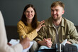 © pressmaster - Caucasian young adult woman and Caucasian young adult man sitting together holding hands smiling while attending counseling session with professional writing on clipboard