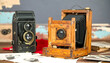 © ArtNote - Two antique cameras, a black and a light brown wooden one, sit on a table with old photographs.