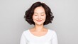 © Neuro architect - Asian woman with curly hair, smiling gently with closed eyes, wearing a white shirt, against a soft gray background, radiating calmness and serenity in a peaceful moment