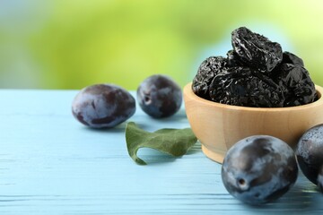 Wall Mural - Dried prunes in bowl, leaf and fresh plums on light blue wooden table, closeup. Space for text