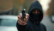 © miss irine - Man in black hoodie, face mask points handgun directly at camera creating tense atmosphere. Close-up urban street scene with blurred background. Fear, intimidation conveyed through confrontation,